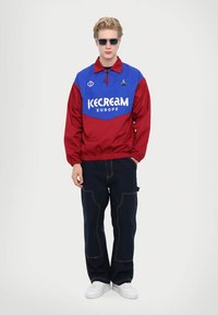 Giacca a pullover rossa e blu con logo "ICECREAM EUROPE" e colletto a contrasto. Indossata con pantaloni blu navy e sneakers bianche.