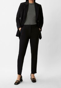 Czarna, dopasowana marynarka na szarym topie w paski, zestawiona z czarnymi, dopasowanymi spodniami i mokasynami na obcasie z detalem w postaci złotego łańcucha.