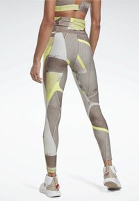 Person som bär högmidjade, mönstrade leggings med beige, vit och gula abstrakta former, kombinerat med en beige sport-bh och vita sneakers.