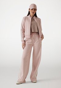 Ensemble de survêtement rose comprenant une veste zippée avec des manches rayées, un pantalon large assorti et une casquette coordonnée avec un détail logo.