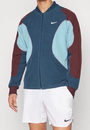Uomo che indossa una giacca Nike blu, azzurra e bordeaux con zip e pantaloncini bianchi Nike, in piedi con una mano parzialmente nella tasca.