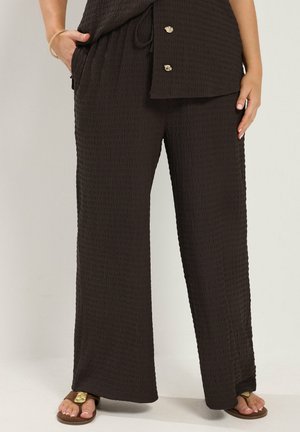 Pantalon large noir texturé porté avec un haut assorti et des sandales marron, les mains reposant sur les hanches et une poche avec un bracelet visible.