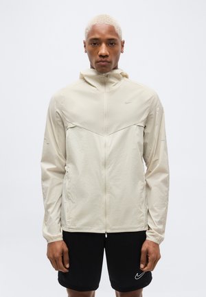 Άτομο με κοντά ξανθά μαλλιά φοράει μπεζ ζακέτα με φερμουάρ και μαύρο σορτς με λευκό λογότυπο Nike, σε απλό ανοιχτό φόντο.