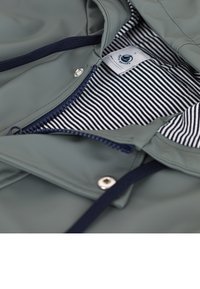 Regenjas in gedempt groen, met een marineblauwe rits en trekkoorden. Het interieur heeft een gestreepte zwart-witte voering met een logo label.
