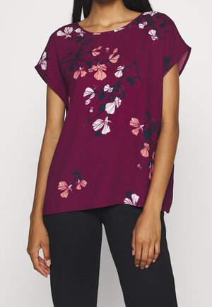 Vrouw draagt een paarse blouse met korte mouwen met roze en witte bloemenprint en zwarte broek, getoond van schouders tot heupen.