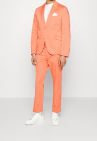 Korallenfarbener Anzug mit einem einreihigen Blazer und passenden Hosen, kombiniert mit einem weißen T-Shirt und weißen Sneakers. Auffälliges Taschentuch-Detail.