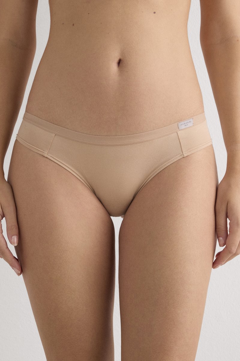 Woman wearing nude seamless bikini-style underwear, standing against a plain light background, showing midsection and upper thighs.