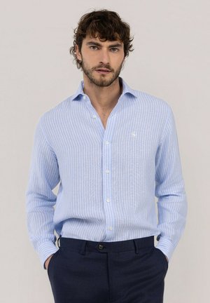 Hombre con cabello rizado que lleva una camisa de rayas azul claro abotonada y pantalones azul oscuro con las manos en los bolsillos.