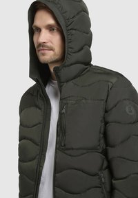 Gefütterte Kapuzenjacke in dunkelgrün. Gestepptes Muster, Reißverschluss, Fronttasche und gesticktes Logo am Ärmel. Leichtes Material.