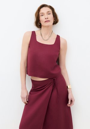 Frau in ärmellosem burgunderfarbenem Top und passendem Wickelrock, dazu goldene Halskette, Creolen und Handgelenkmanschette, stehend.