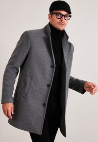 Abrigo de mezcla de lana gris con un corte entallado, cuello con muesca, botones en el frente y bolsillos laterales, combinado con un suéter negro de cuello alto.