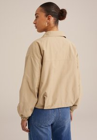 Beige oversized bunda s límečkem, elastickými manžetami a hladkým matným povrchem, kombinovaná s vysokými modrými džíny.
