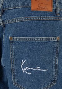 Mėlynos džinsinės kelnės su odiniu ženkleliu su tekstu ir siuvinėtu baltu parašu ant užpakalinio kišelio. Klasikinis penkių kišenių dizainas.