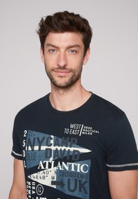 Camiseta de algodón azul oscuro con texto gráfico y diseños náuticos en blanco y azul claro. Mangas cortas con detalle de costura en contraste.