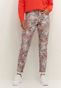 Person iført mønstrede slim-fit bukser med blomsterdesign, en orange sweater, hvide sneakers, stående med hænderne i lommerne.