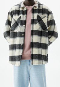 Persoon in een zwart-wit-grijs geblokte button-up jack over een roze trui met lichtblauwe spijkerbroek, staande tegen een effen achtergrond.