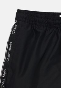 Sorte idrettsshorts laget av glatt stoff, med elastisk midje og sidestriper med "Calvin Klein"-logo i hvitt.