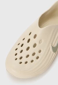 Chaussure légère beige avec de multiples trous de ventilation circulaires, texture lisse, aspérités surélevées et un accent logo métallique sur le côté.