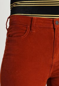 Pantalon en velours côtelé couleur rouille avec poches avant et passants pour ceinture, porté avec une chemise rayée noire et jaune.