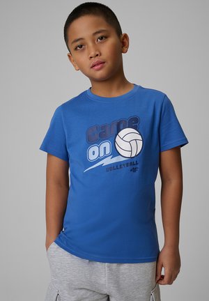 Młody chłopiec ubrany w niebieską koszulkę z napisem "Game On Volleyball" i jasnoszorty, stojący z jedną ręką w kieszeni na jednolitym tle.