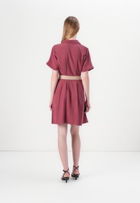 Robe courte bordeaux avec des manches courtes et un col, cintrée à la taille par une ceinture tissée. Tissu lisse et longueur au genou.