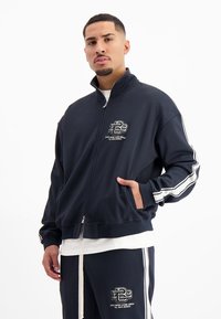 Marineblauwe zip-up jas met een hoge kraag, witte streepaccenten op de mouwen en voorzakken. Heeft een logo op de borst. Zachte stoftextuur.