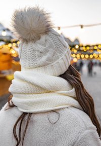 Knitted white hat with a faux fur pom-pom on top, paired with a thick, textured white scarf, set against a blurred festive backdrop.