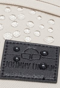 Beige textúrájú anyag, amelyen tiszta vízcseppek láthatók, fekete bőr címkével, amelyen a "JOHNNY URBAN" felirat és egy szemüveg ikon látható.