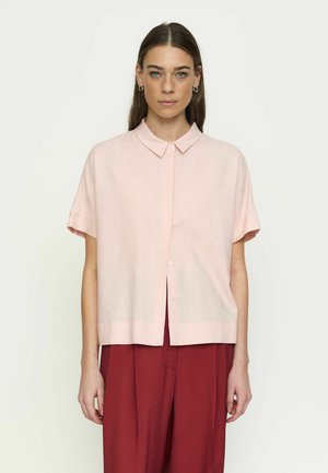 Kobieta o długich brązowych włosach, ubrana w jasnoróżową koszulę na krótkie rękawy zapinaną na guziki oraz czerwone luźne spodnie, stojąca na jednolitym jasnym tle.