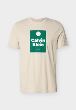 Beigen värinen lyhythihainen t-paita, jossa vihreä suorakulmainen logo, jossa on valkoinen ympyrä ja "Calvin Klein" -teksti paksuilla valkoisilla kirjaimilla.