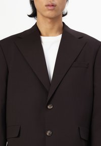 Tummanruskea bleiseri, joka on valmistettu sileästä kankaasta ja jossa on nappikaulus, yksi rintatasku ja kaksi nappia. Päällä valkoinen paita.