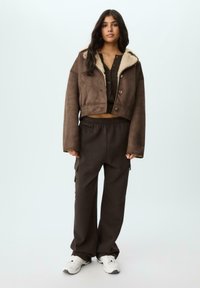 Giacca corta marrone con fodera in pelliccia sintetica e chiusura a bottone, abbinata a joggers marroni larghi con tasche laterali e sneakers bianche.