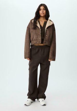 Braune, verkürzte Jacke mit Schafsfutter und Knopfverschluss, kombiniert mit lockeren braunen Jogginghosen mit Seitentaschen und weißen Sneakers.