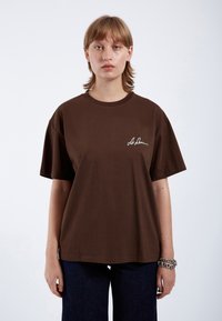 Brown cotton T-shirt with short sleeves, round neck, and white embroidered logo on the left chest. Worn with dark blue denim pants.