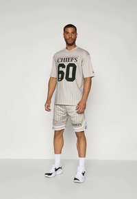 Camisola atlética cinzenta com "CHIEFS" e "60" impressos a preto; combinada com calções a condizer com riscas e detalhes de logótipo.