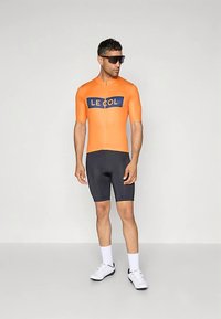 Πορτοκαλί ποδηλατική φανέλα με κοντά μανίκια και εμπρόσθιο φερμουάρ, με μπλε λογότυπο. Μαύρο στενό σορτς και λευκά ποδηλατικά παπούτσια.