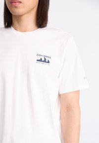 White cotton t-shirt featuring a navy printed logo on the left chest and a small accent on the sleeve. Short sleeves, crew neck design.