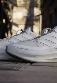 Scarpe da corsa bianche Adidas con testo sulla suola "Light Boost", posizionate su una strada lastricata con edifici sfocati sullo sfondo.