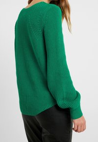 Zelený pletený svetr s žebrovanou texturou, dlouhými rukávy a zaobleným výstřihem, doplněný tmavými koženými kalhotami.