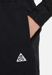 Juodi elastiniai kelnaičių diržai su šonine kišenėle ir baltu trikampiu logotipu, išsiuvinėtu apačioje, pasižymi lygiu tekstūra ir lengvu audiniu.