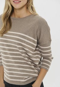 Donna che indossa un maglione beige e bianco a righe orizzontali con maniche a tre quarti, in piedi con una mano nella tasca dei pantaloni neri.