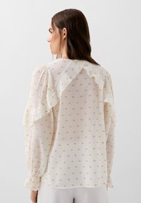Blouse blanche avec un dos en volants et des manches longues, présentant un tissu texturé avec des motifs carrés dorés.