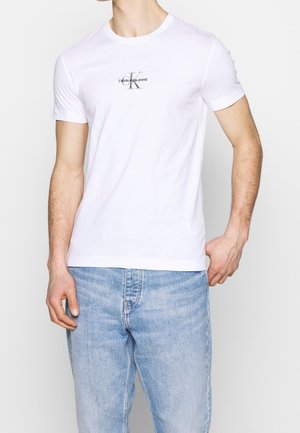 Mand iført en hvid Calvin Klein Jeans t-shirt og lyseblå denimjeans, stående med den ene hånd i lommen på en ensfarvet baggrund.