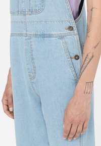 Salopette in denim azzurro chiaro con vestibilità rilassata, dotata di accessori in argento, bottoni laterali e tasca sul petto. Dettaglio di texture verticale sottile.