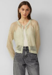 Mintgrüner Netzcardigan mit rundem Halsausschnitt, langen Ärmeln, Knopfverschluss, gerippten Bündchen und aufwendig gestricktem Muster. Kombiniert mit grauen Jeans.