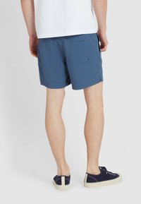 Blaue Shorts aus strukturiertem Stoff mit einer Gesäßtasche und Seitenschlitzen. Kombiniert mit dunkelblauen Sneakers und einem weißen Shirt.