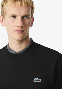 Sudadera negra de algodón con cuello redondo, que presenta una camisa de color gris debajo y un logo de cocodrilo de colores en el pecho.