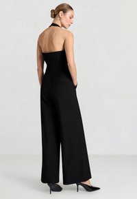 Donna che indossa una tuta nera senza spalline a collo alto con gambe larghe e scarpe nere con tacco alto, in piedi contro uno sfondo chiaro e uniforme.