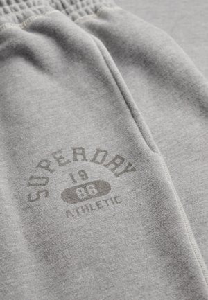 Pantalones de chándal gris claro con cintura elástica y un logotipo impreso que dice "Superdry 1986 Athletic" cerca del bolsillo.