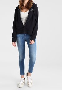Felpa blu navy con zip e cordini, abbinata a jeans skinny azzurri e sneakers bianche con dettagli floreali.